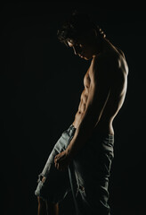 Fototapeta premium Young man stands shirtless in shadows, highlighting muscular physique. Wearing ripped jeans, the image conveys strength, fitness, and confidence.