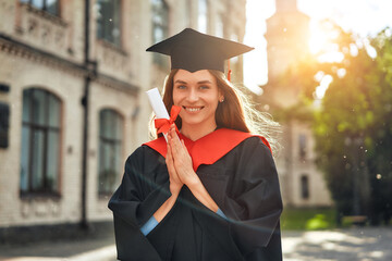 A happy graduate holding a diploma in front of a university building on a sunny day
