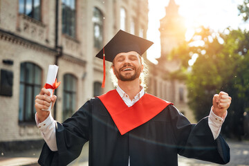 A male graduate is celebrating his academic achievement with a diploma outdoors on a sunny day