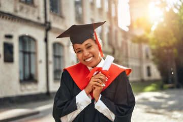 A happy graduate holding a diploma in front of a university building on a sunny day