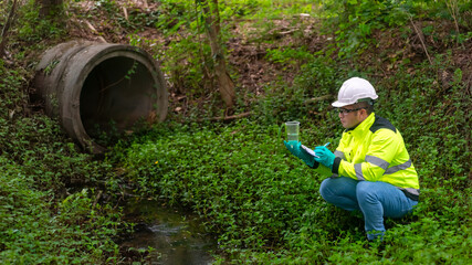 An ecologist in a green jacket sit aside of the pond looking at a liquid in a plastic glass in his...