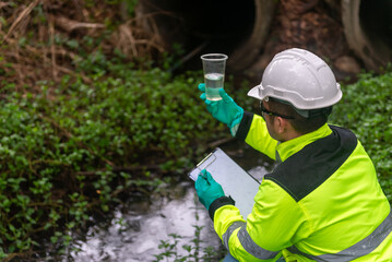 An ecologist in a green jacket sit aside of the pond looking at a liquid in a plastic glass in his hand try to examination the quality of the water. and write down the result on a clipboard