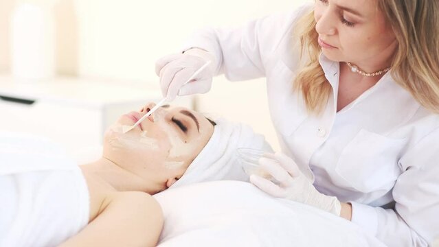 professional cosmetologist applies a white mask on the face of a female client with a brush, close-up