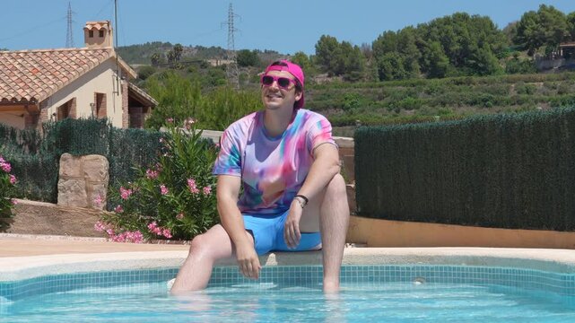 A man in a colorful shirt and blue shorts sits in a pool. He is wearing sunglasses and a pink hat