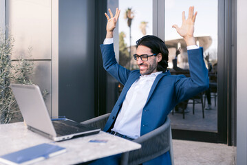 A young businessman in a suit and glasses sitting outdoors with laptop celebrating success with arms raised