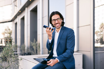 smiling Caucasian businessman with digital tablet talking by cellphone using white wireless earphones while sitting on street near modern office building