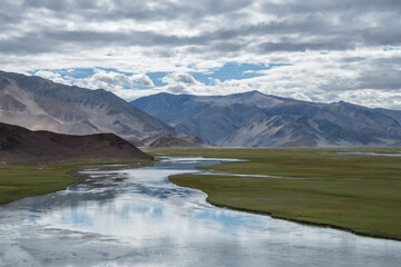 A river flows through a grassy field with distant mountains, Ladakh, Himalayas