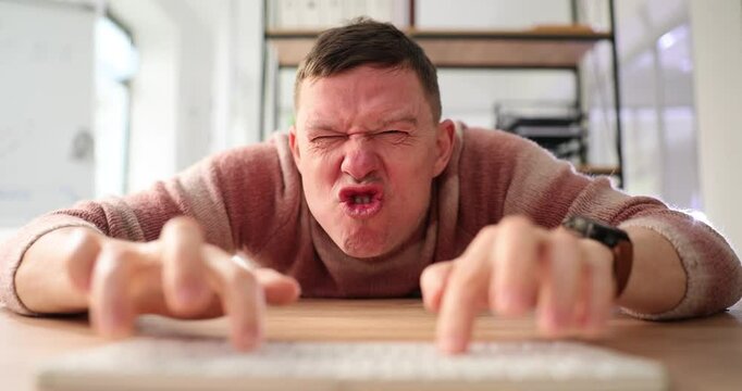Excited grimacing internet fraud guy types on PC keyboard at table slow motion. Crazy man prepares provocation online. Emotional gamer plays in room