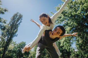 A young man gives a piggyback ride to a woman in a park, both are laughing joyfully with arms...