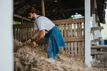 Man in a barn handling hay, wearing a white shirt, jeans, hat, and sunglasses. Rural and farm...
