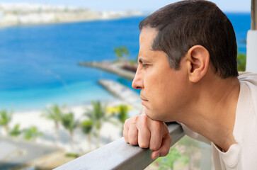 Side view portrait of a sad single man looking down from a balcony of a hotel with an beach background