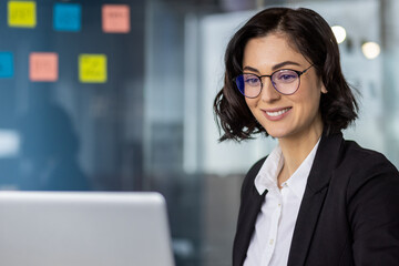 Confident businesswoman with glasses smiling while working on laptop in a modern office environment. Professional and focused on her tasks.