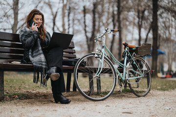 Elegant young woman in stylish attire engrossed in work on her laptop while sitting in a city park.