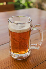Glass mug with fresh cold beer on wooden table on patio. A refreshing summer drink. Liquor. A pint of beer. Foam beer in a glass mug. Street cafe. Summer vacation. Abnormal heat.