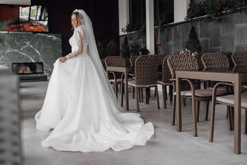 A bride in a white dress stands in front of a fireplace. She is wearing a veil and a long white gown. The scene is set in a restaurant with several chairs and tables