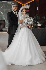Fototapeta premium A bride and groom are posing for a picture in front of a green plant. The bride is wearing a white dress and the groom is wearing a suit. The bride is holding a bouquet of flowers