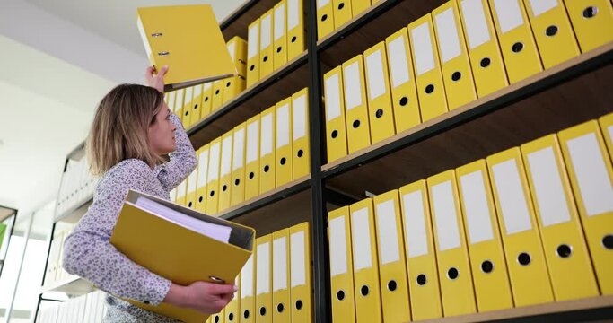 Woman reaches hand to get yellow folder on upper shelf of rack in office. Secretary looks for ring binder with annual accounting materials for paperwork slow motion