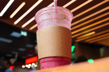 A plastic cup filled with a pink beverage sits on a table in a fast food restaurant, against a blurred background of ceiling lights, a low-angle shot. Paper cup sleeve mockup.