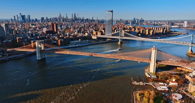 Yachts sail under the Manhattan and Broadway Bridges at sunset. Drone footage distancing from the bridges revealing the vast scenery of metropolis.