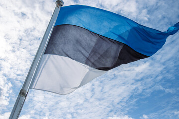 Beautiful flag of Estonia waving in the wind on the flagpole against blue sky with clouds. Close up of Estonian national flag fluttering on windy day. Cloudy sky background. Copy space.