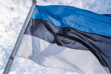 Beautiful flag of Estonia waving in the wind on the flagpole against blue sky with clouds. Close up of Estonian national flag fluttering on windy day. Cloudy sky background. Copy space.