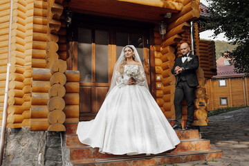Fototapeta premium A bride and groom stand outside a wooden house, the bride wearing a white dress and the groom wearing a suit