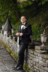 A man in a black suit and tie stands in front of a stone wall. He is wearing a white tie and a boutonniere