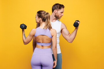 Back rear view young strong fitness trainer instructor sporty two man woman wearing blue clothes spend time in home gym hold dumbbells isolated on plain yellow background. Workout sport fit concept.