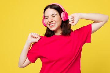 Young smiling happy cheerful woman wear pink t-shirt casual clothes listen to music in headphones raise up hands dance isolated on plain yellow orange background studio portrait. Lifestyle concept.