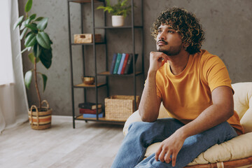 Young minded pensive thoughtful Indian man he wear orange casual clothes prop up chin sits in armchair stay at home hotel flat rest relax spend free spare time in living room indoor. Lounge concept.