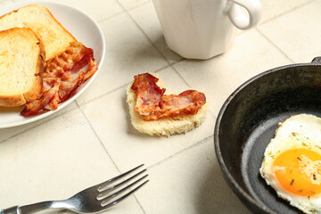 Frying pan with tasty fried egg, toasts and bacon on light tile background, closeup