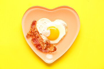 Plate with tasty bacon and heart made of fried egg on yellow background