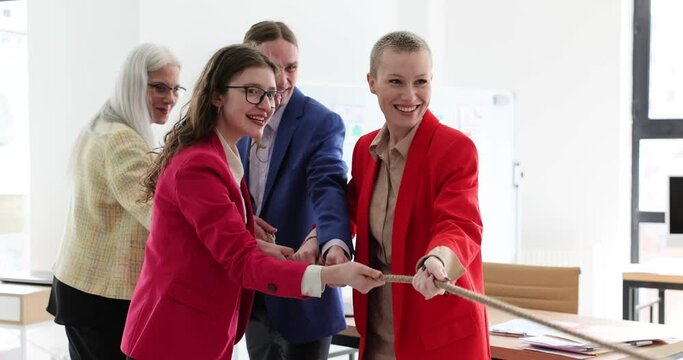 Happy colleagues in colorful jackets pull rope together and have fun at team-building. Delighted collective develops teamwork skills in spacious light office