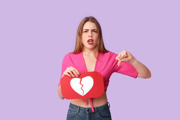 Beautiful young displeased woman with heart dislike icon showing thumb-down gesture on purple...