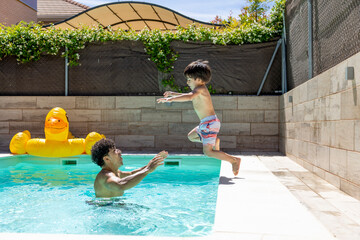 Latin father catching son jumping into pool, yellow duck float in background, sunny day.