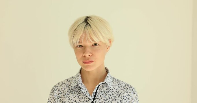 Stylish young woman in patterned office blouse winks and looks skeptically. Stylish woman with short blonde hairdo expresses mistrust against beige wall