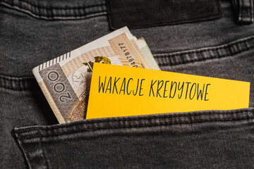 Yellow card with a handwritten inscription "Wakacje Kredytowe", inserted into the pocket of gray pants jeasnow, next to Polish banknotes PLN (selective focus), translation: Credit Holidays