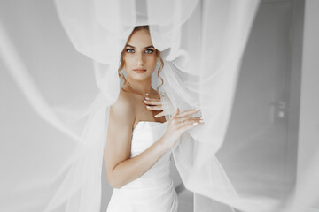 A woman in a white wedding dress is standing in front of a curtain. She is wearing a veil and has...