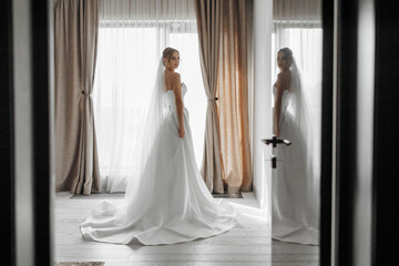 A bride is standing in front of a window in a room with curtains. She is wearing a white dress and...