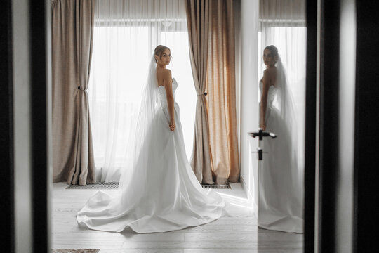 A bride is standing in front of a window in a room with curtains. She is wearing a white dress and a veil