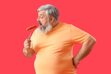 Mature man eating tasty sausage on red background
