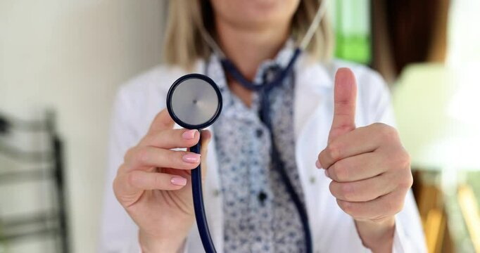 Pleased doctor holds stethoscope in hand showing thumb-up. Woman approves serviceability of medical equipment standing in clinic office slow motion