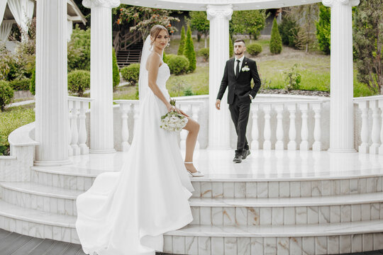 A bride and groom are standing on a marble staircase in front of a gazebo. The bride is holding a bouquet and the groom is wearing a suit. The scene is elegant and romantic - Powered by Adobe
