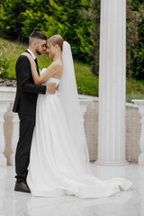 Naklejka premium A bride and groom are standing together, embracing each other. The bride is wearing a white dress and the groom is wearing a black suit. The scene is set in a garden
