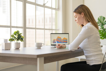 Young woman at desk next to a large window and writing notes