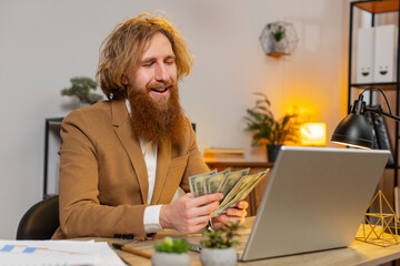 Happy Caucasian bearded businessman counting money cash and using laptop in home office. Successful...