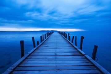 Obraz premium A dramatic pier stretches out into the brightly lit blue sea beneath a sweeping, clouded sky, conveying both the beauty and dynamism of coastal and seascape photography.