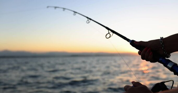 Female hand holds fishing rod. Woman fishes standing on sailing motorboat at summer sunset. Active recreation and hobby slow motion