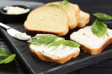 Pieces of bread with cream cheese and basil leaves on black table, closeup