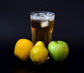 Apple and pear juice with ice in a tall faceted glass on a black background, with ripe fruits nearby.